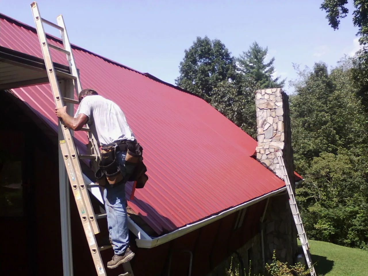 Roofer on a ladder beside a red metal roof and stone chimney — Soffit & Fascia Repair by Roofing Brothers in Rancho Palos Verdes